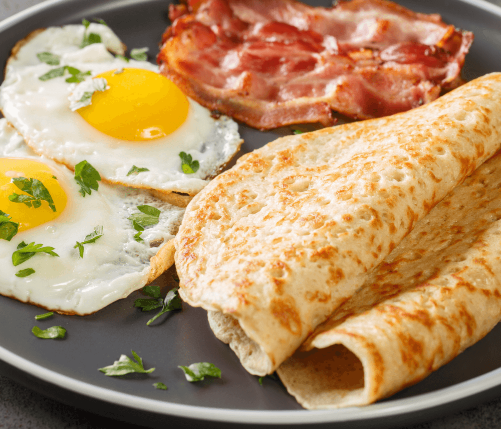 A plate with two fried eggs, two slices of crispy bacon, and two folded crepes with parsley garnish. - Home Instead