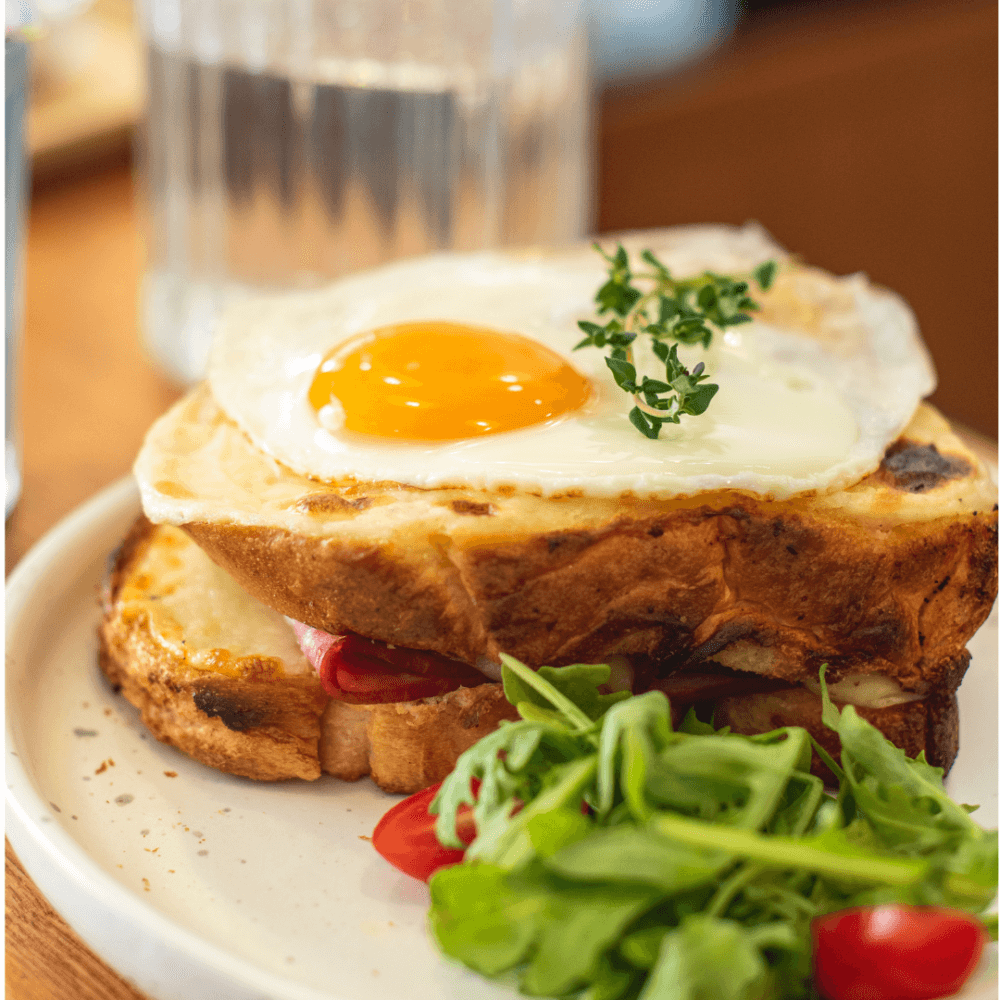 A fried egg on toast with greens and cherry tomatoes on a white plate. A glass of water is in the background. - Home Instead