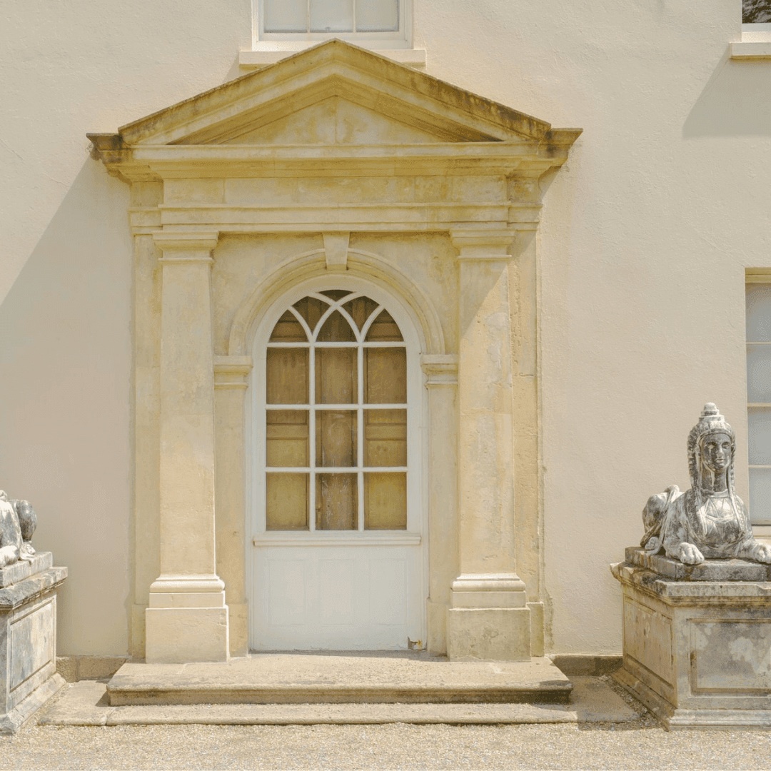 A classical arched doorway with stone sculptures and a decorative pediment on a pale exterior wall. - Home Instead