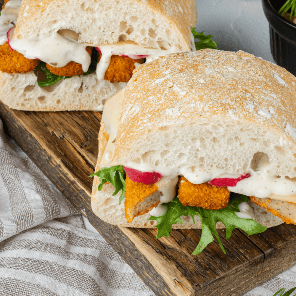 Two crispy chicken sandwiches with lettuce, radish slices, and creamy sauce on ciabatta bread, served on a wooden board. - Home Instead