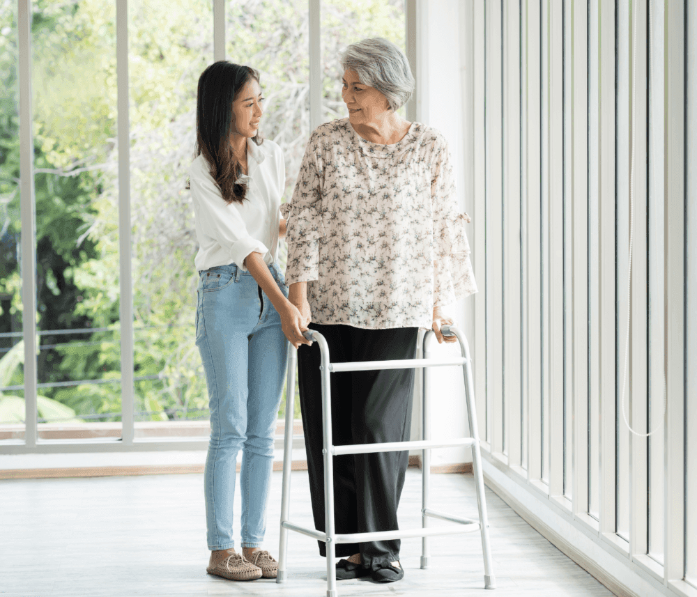 An elderly woman using a walker is assisted by a young woman in a bright room with large windows. - Home Instead