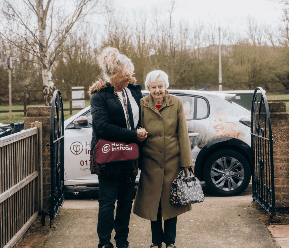 A caregiver escorts an elderly woman through a gate. A car with "Home Instead" branding is parked in the background. - Home Instead