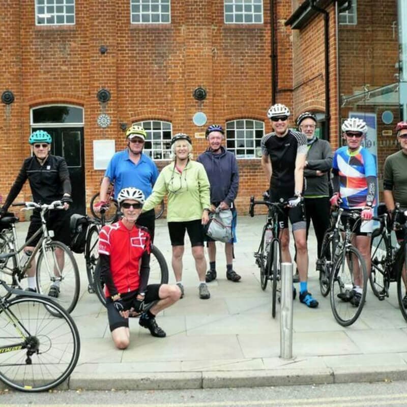 A group of cyclists wearing helmets pose for a photo in front of a brick building with their bicycles. - Home Instead