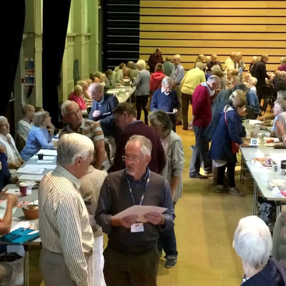 A large group of people gathered in a hall, engaging in conversations and activities around tables. - Home Instead