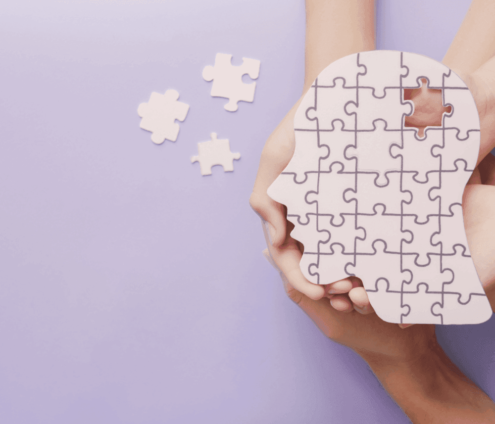 Hands holding a human head-shaped jigsaw puzzle on a purple background, with a few puzzle pieces on the side. - Home Instead