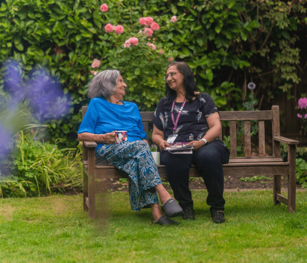 Two women sitting on a wooden bench in a garden, smiling and conversing with each other. - Home Instead
