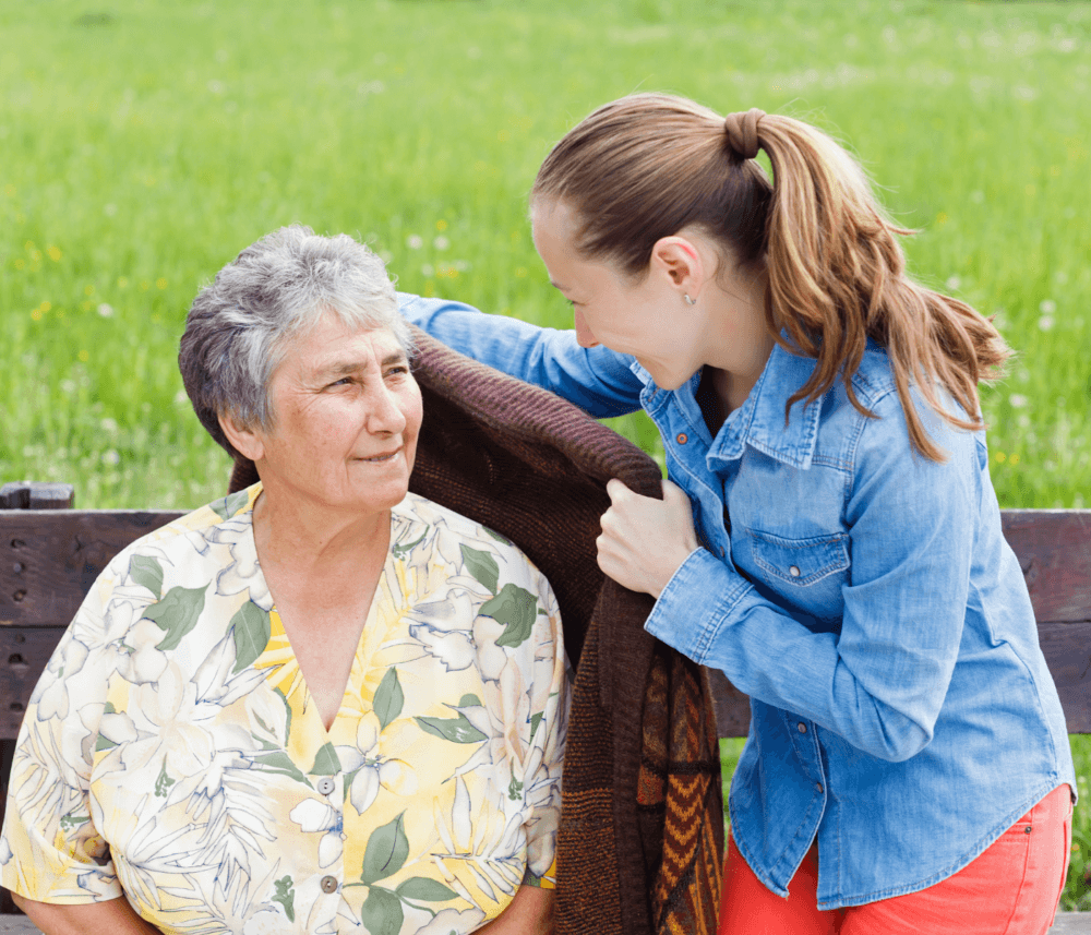 A young woman in a blue shirt places a blanket on an elderly woman sitting on a bench in a grassy field. - Home Instead