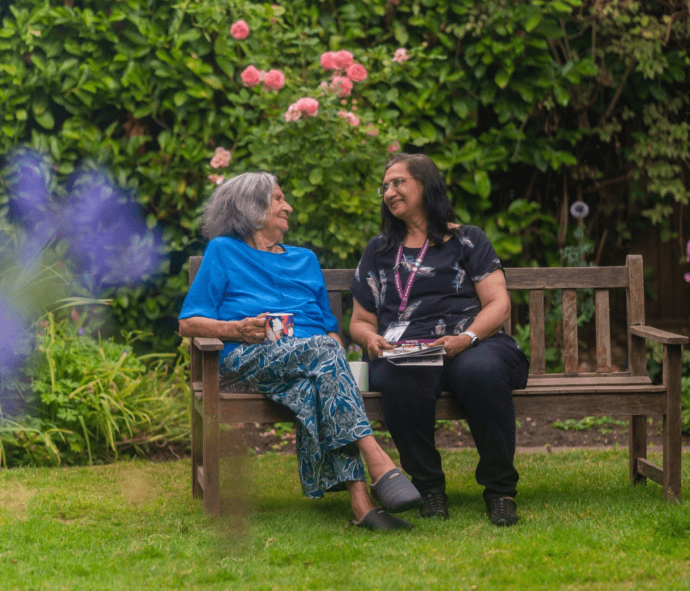 Two women sitting on a wooden bench in a garden, smiling and talking, with flowers and greenery in the background. - Home Instead