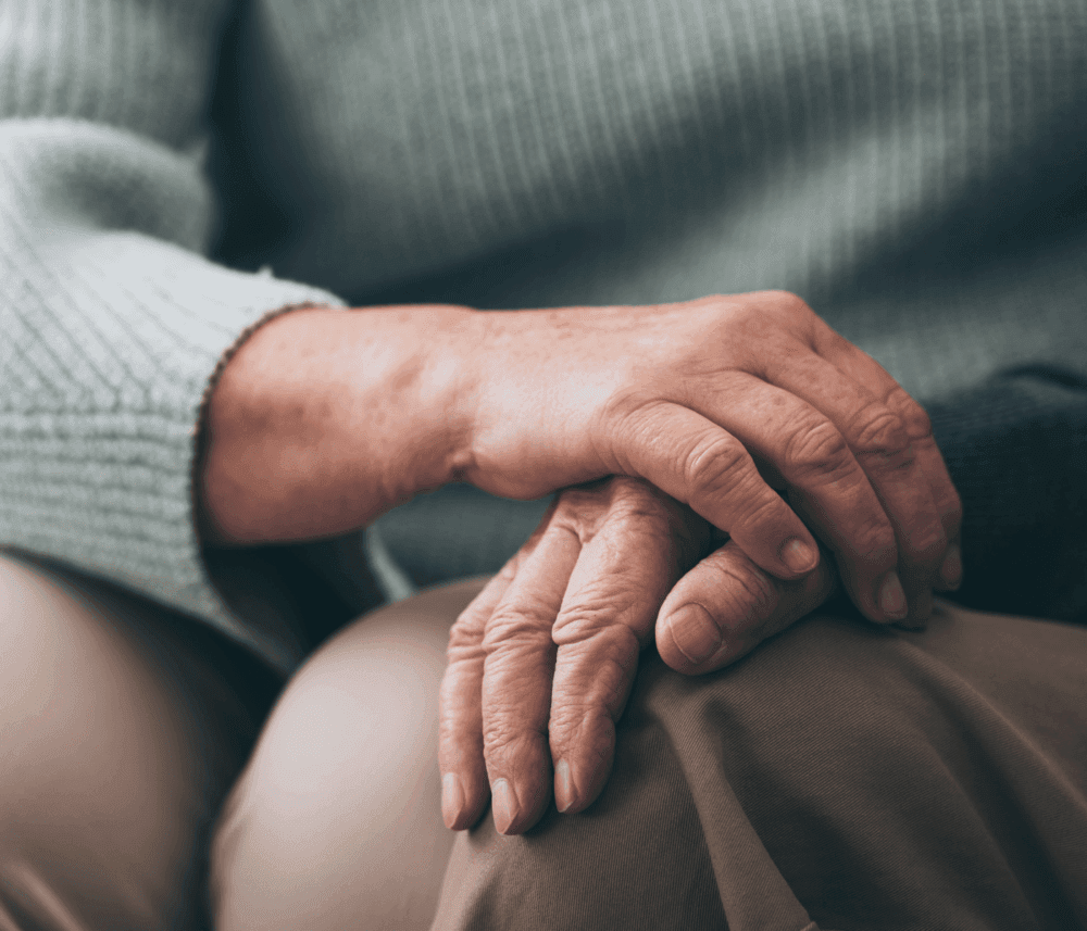 Close-up of an elderly person’s hands resting on their lap, with one hand gently placed over the other. - Home Instead