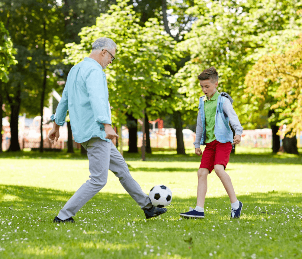 An elderly man and a young boy kicking a soccer ball together in a park on a sunny day. - Home Instead