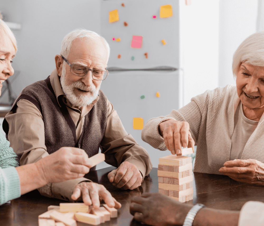 Four elderly people play a game of Jenga, smiling and concentrating around a table in a cozy room. - Home Instead