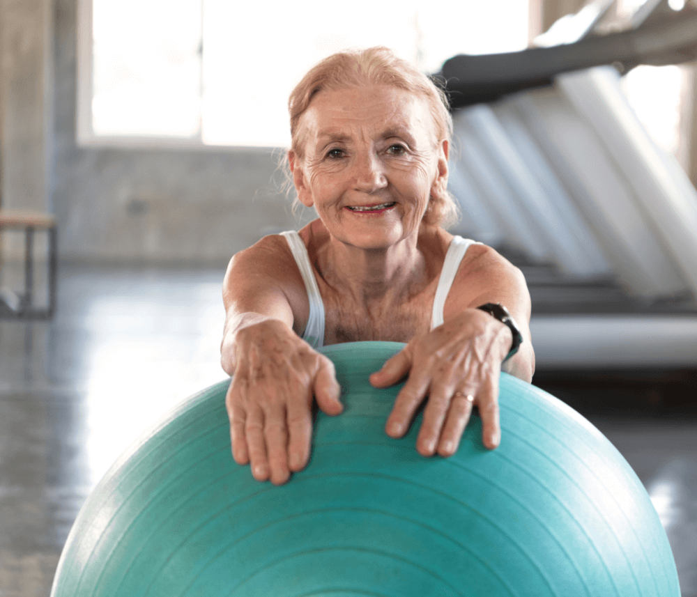 Elderly woman in a gym, smiling and leaning on a blue exercise ball with treadmills in the background. - Home Instead