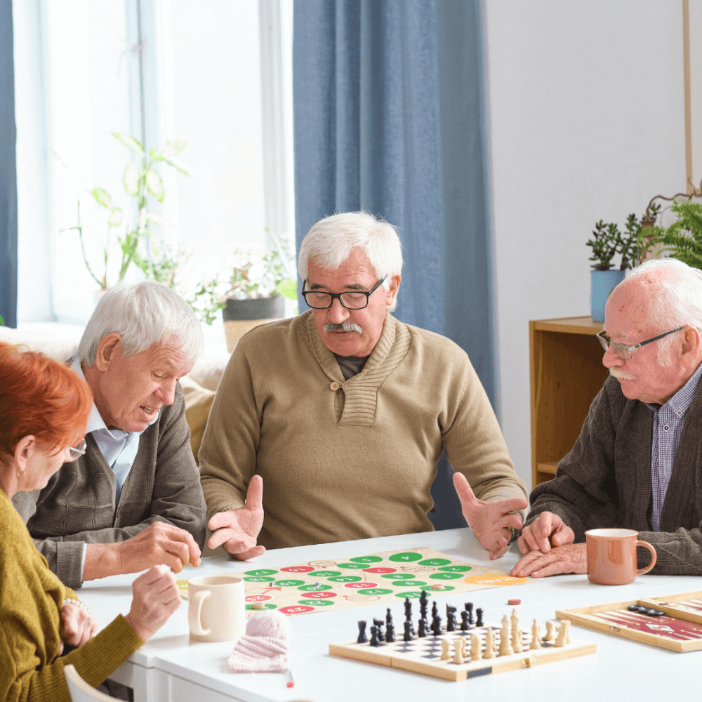 Four elderly people sitting around a table playing board games and puzzles, with coffee mugs and bright natural light. - Home Instead