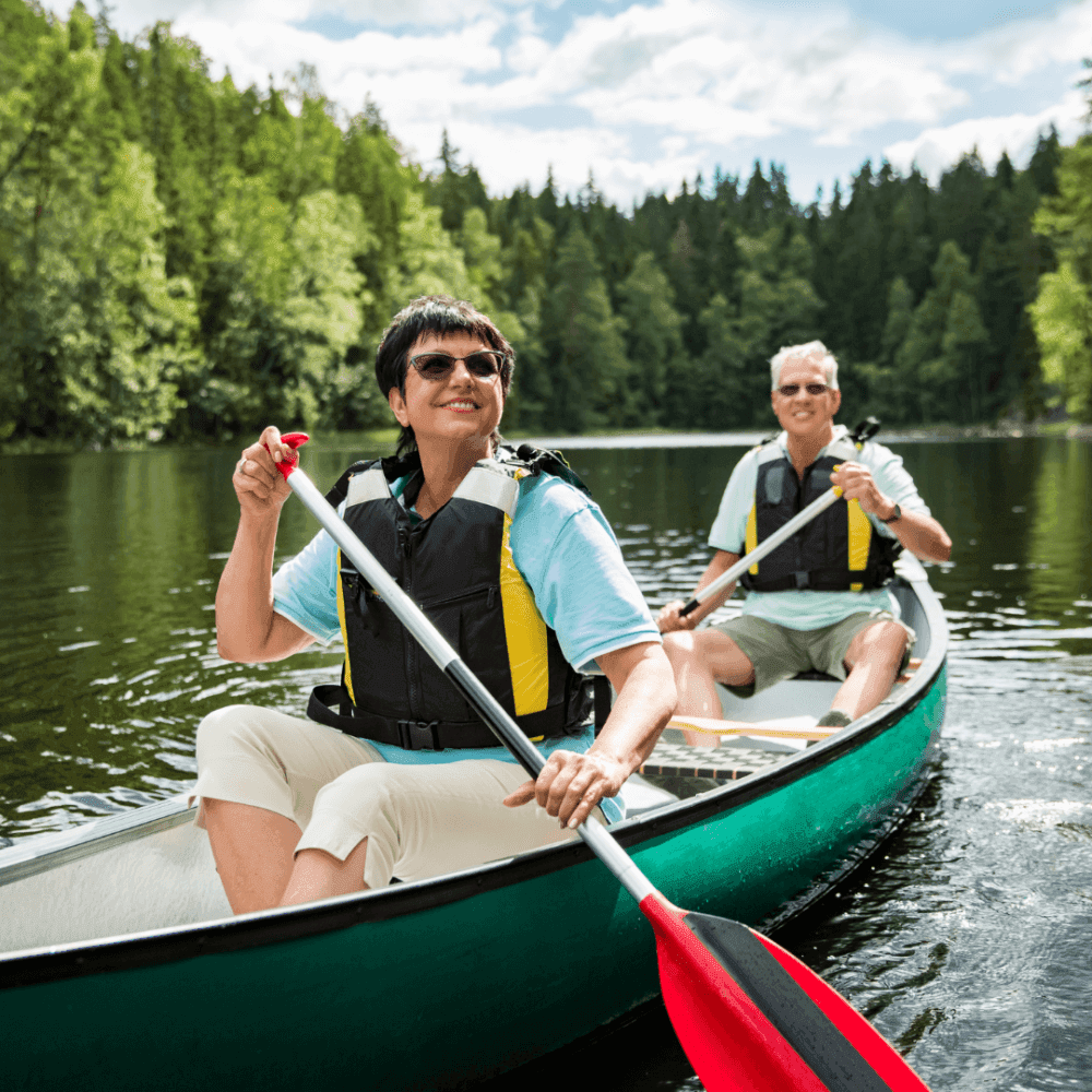 Two people in life jackets paddling a canoe on a peaceful lake surrounded by lush green trees and a cloudy sky. - Home Instead
