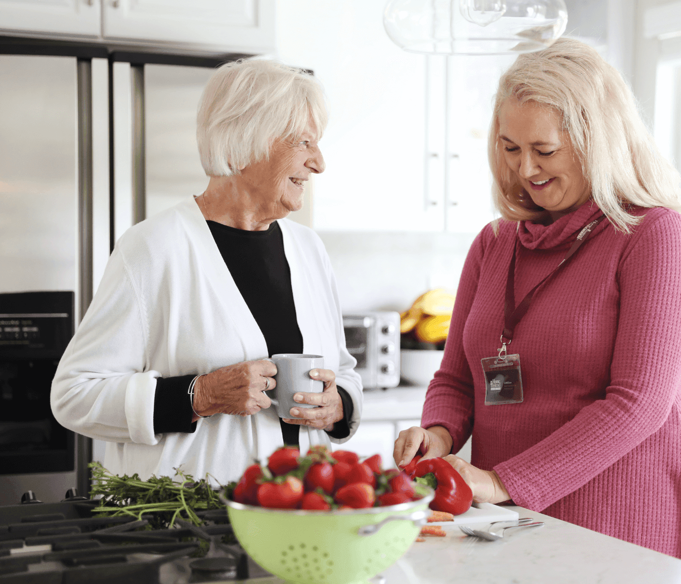 Two women smiling in a kitchen while chopping vegetables, with a bowl of strawberries in the foreground. - Home Instead