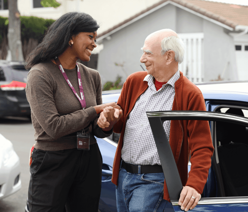 A caregiver helps an elderly man out of a car. They are smiling and holding hands in a neighborhood setting. - Home Instead
