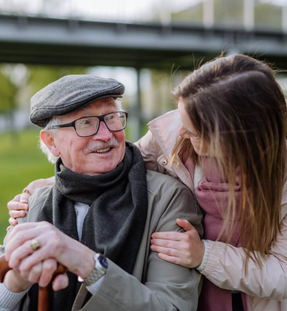 An elderly man smiles, wearing glasses and a cap, while a younger woman lovingly embraces him outdoors. - Home Instead