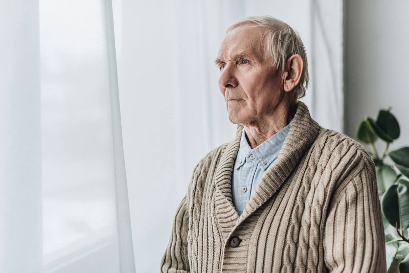 Elderly man in a knitted sweater gazing out of a window beside a potted plant. - Home Instead