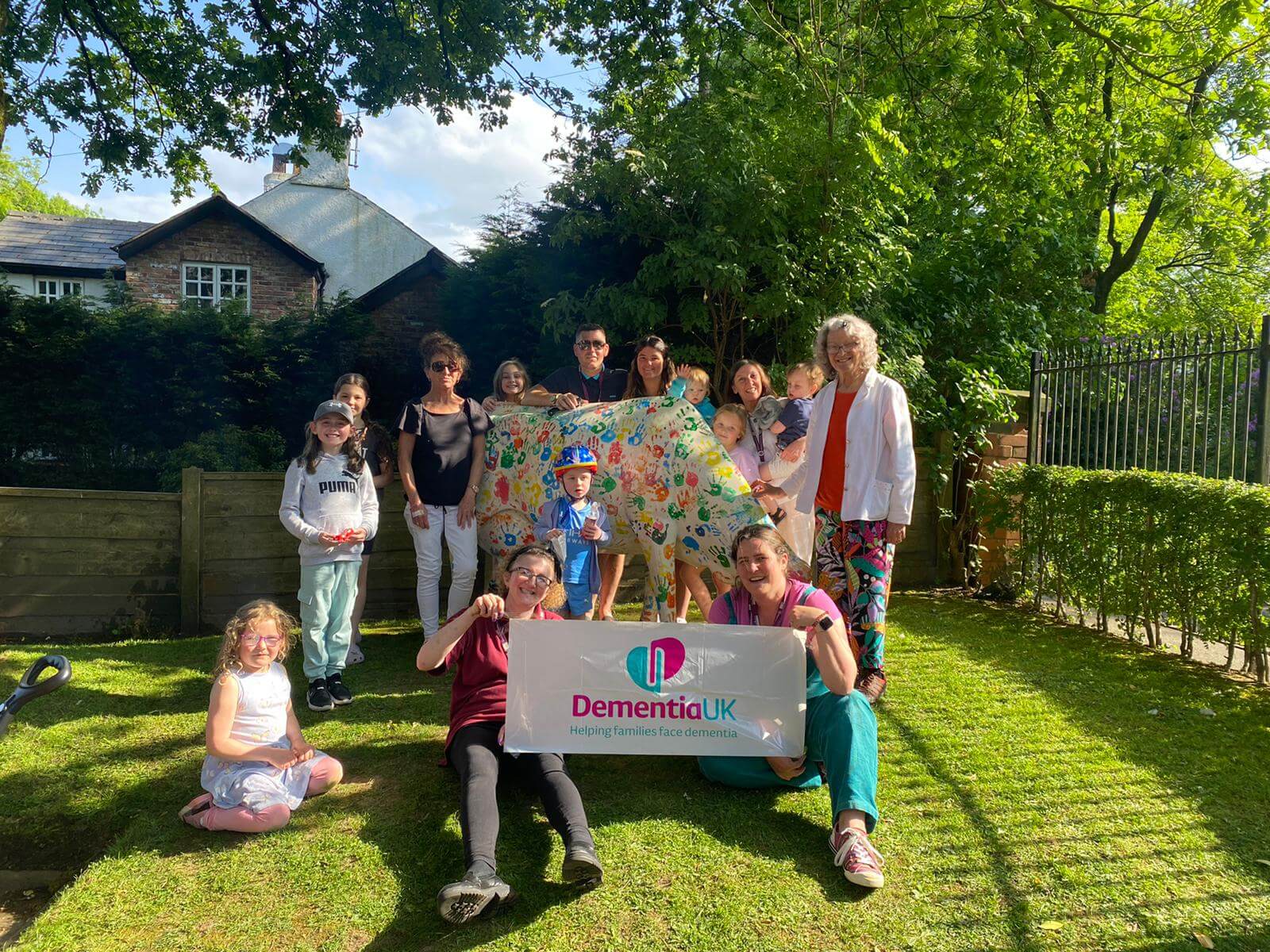 A group of people, including children, holding a colorful banner and a Dementia UK sign, standing outdoors on grass. - Home Instead