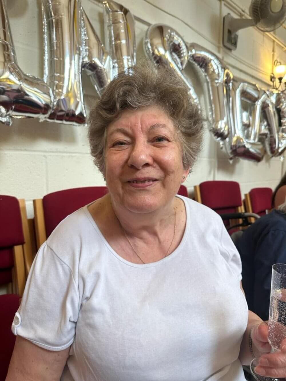 An older woman with short gray hair smiles and holds a glass, with silver balloons in the background that spell "INSPIRATION. - Home Instead