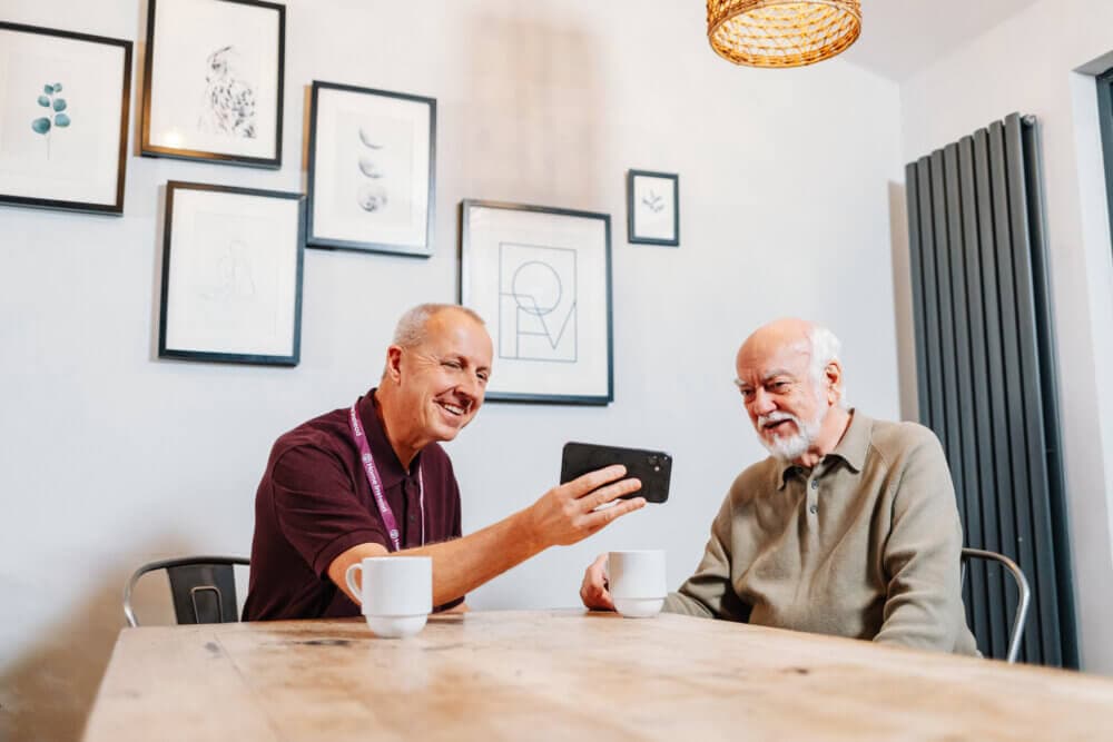 Two men sitting at a wooden table with coffee cups, looking at a smartphone. Artwork decorates the wall behind them. - Home Instead