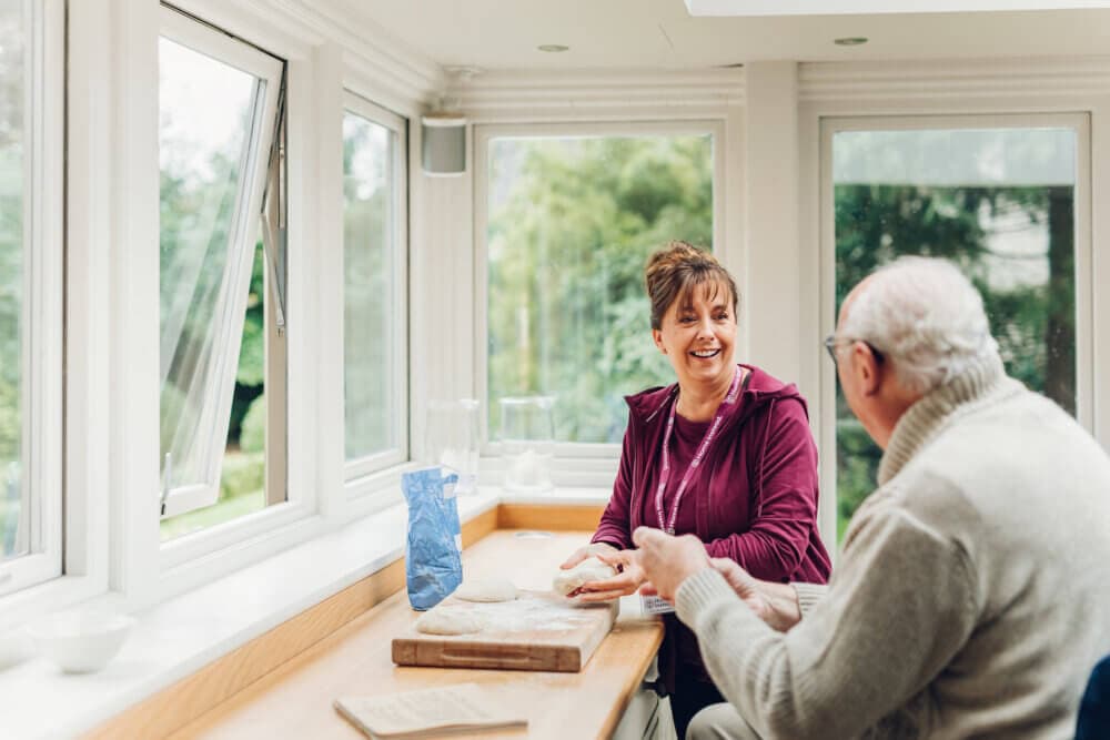 A caregiver and an elderly man are talking and smiling in a bright, window-filled room with greenery outside. - Home Instead