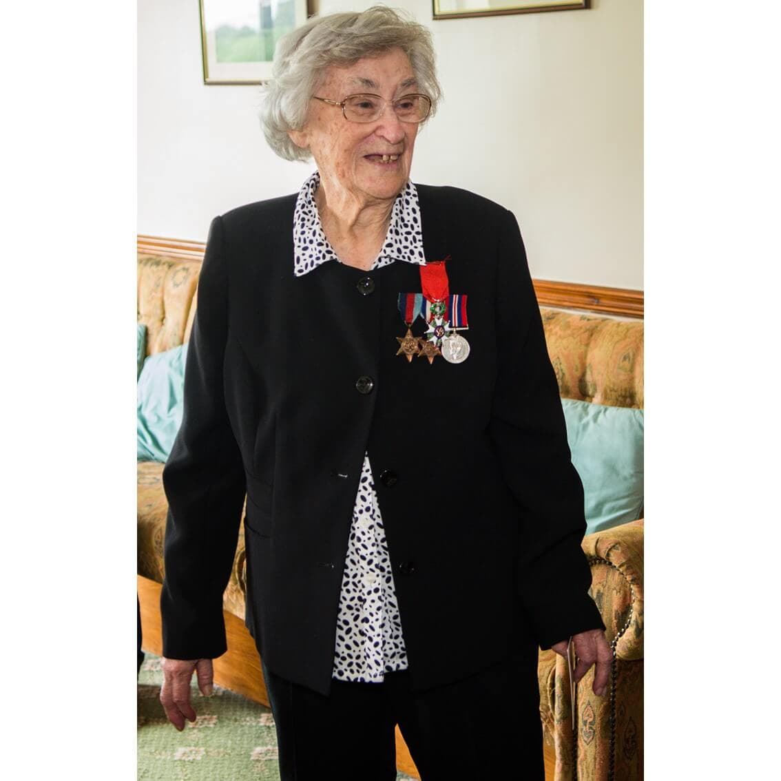 An older woman wearing a black suit with military medals on her chest, standing indoors and smiling. - Home Instead