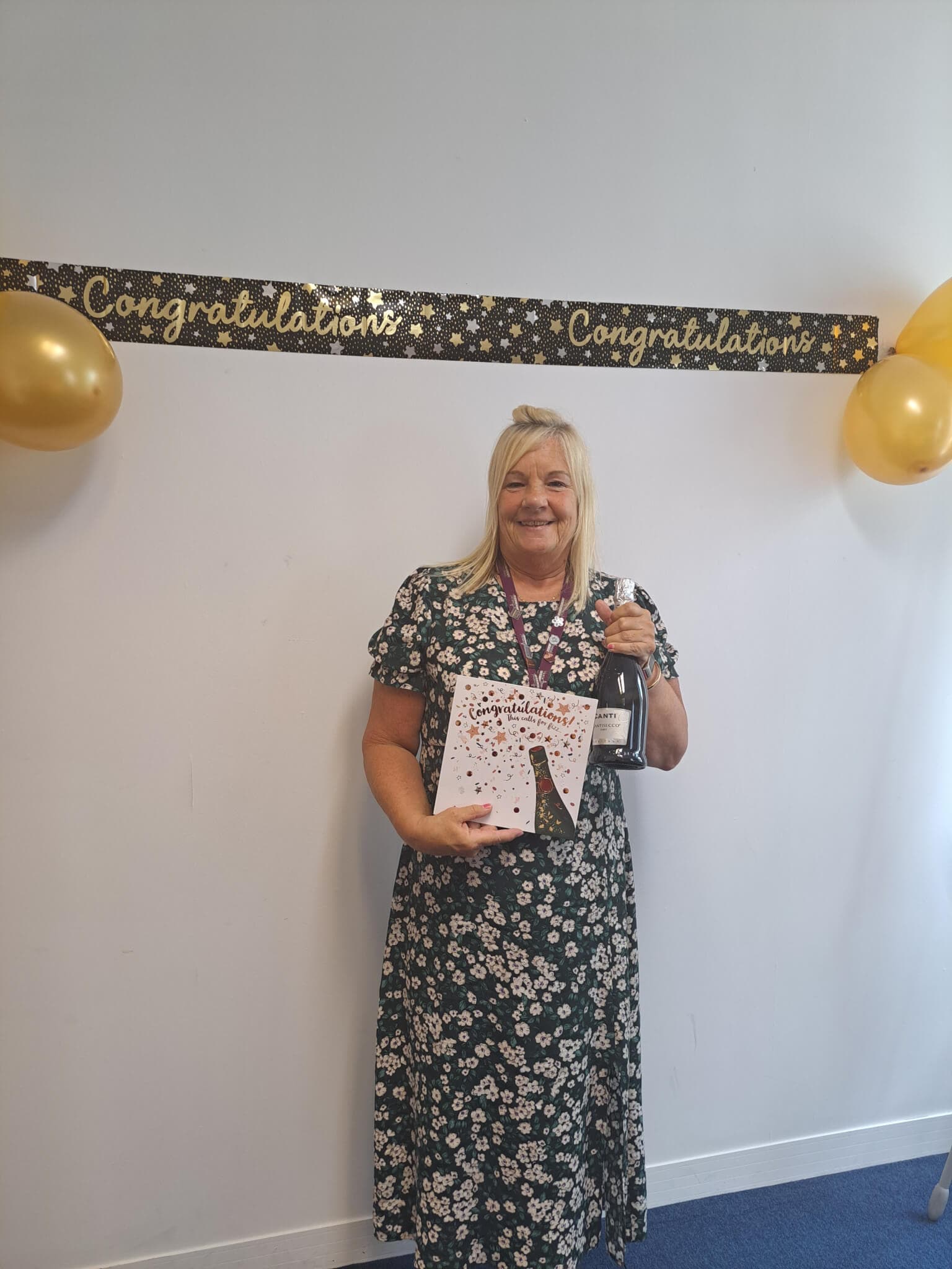 A woman in a floral dress holds a gift and a bottle, standing in front of a "Congratulations" banner with balloons. - Home Instead
