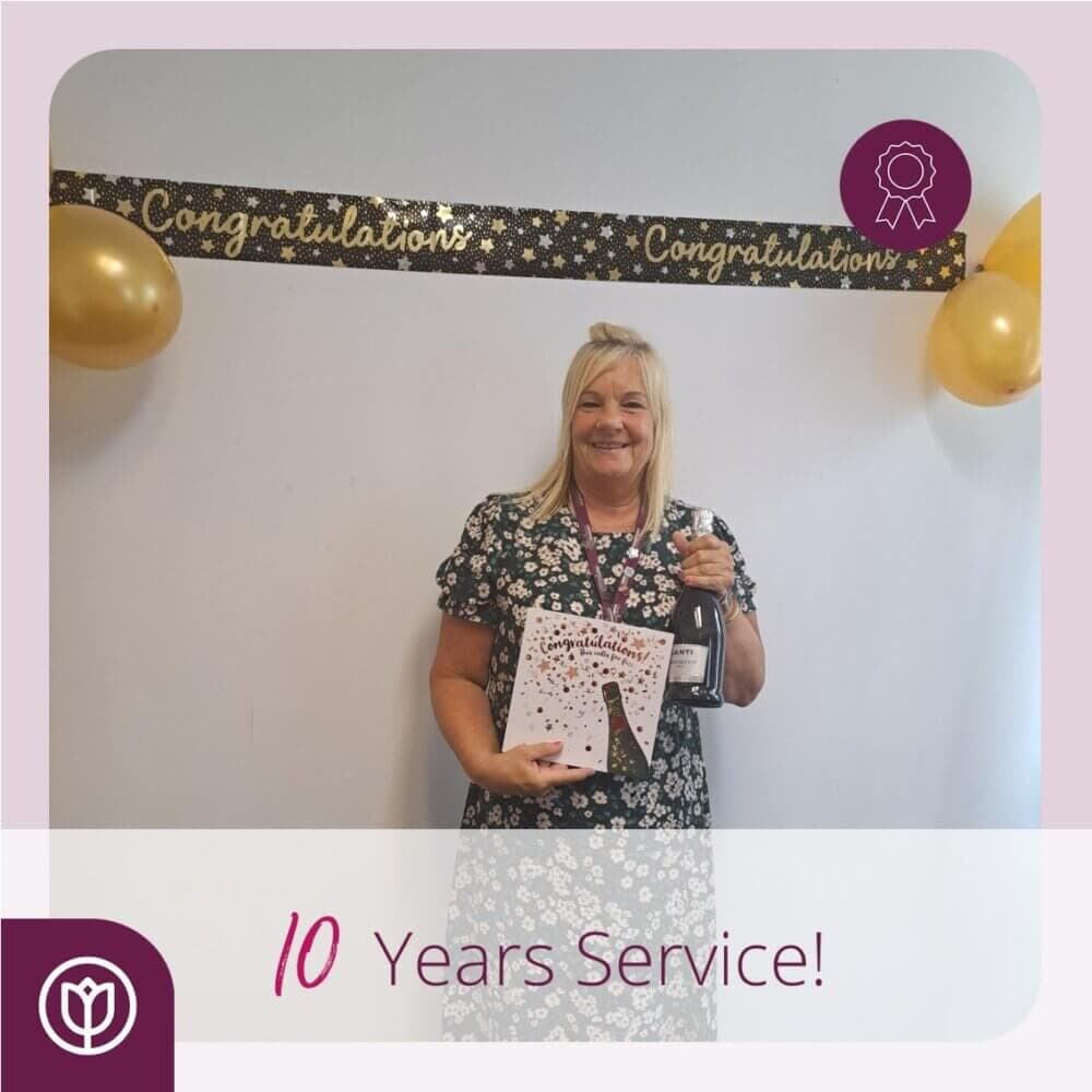 Woman holding a gift and bottle, standing against a decorated wall with "Congratulations." Text reads "10 Years Service!. - Home Instead