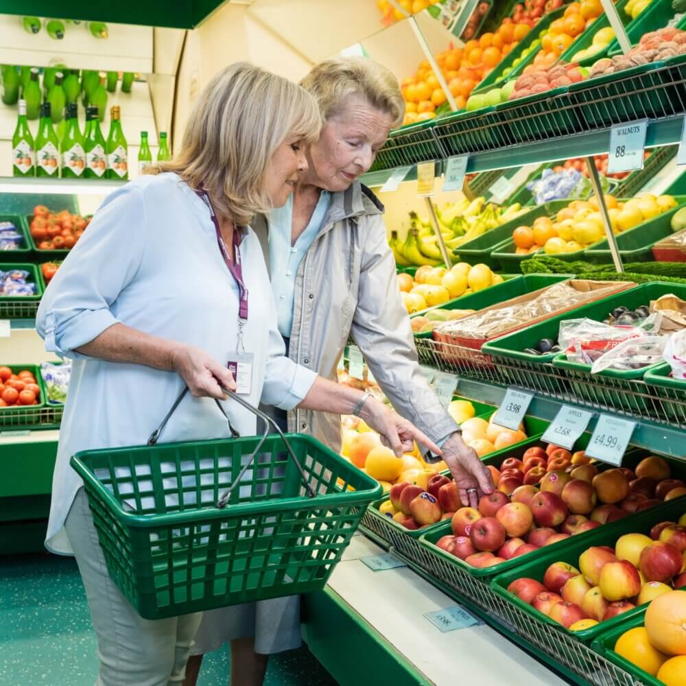 Two women shopping together, selecting apples from a fruit display at a grocery store. - Home Instead
