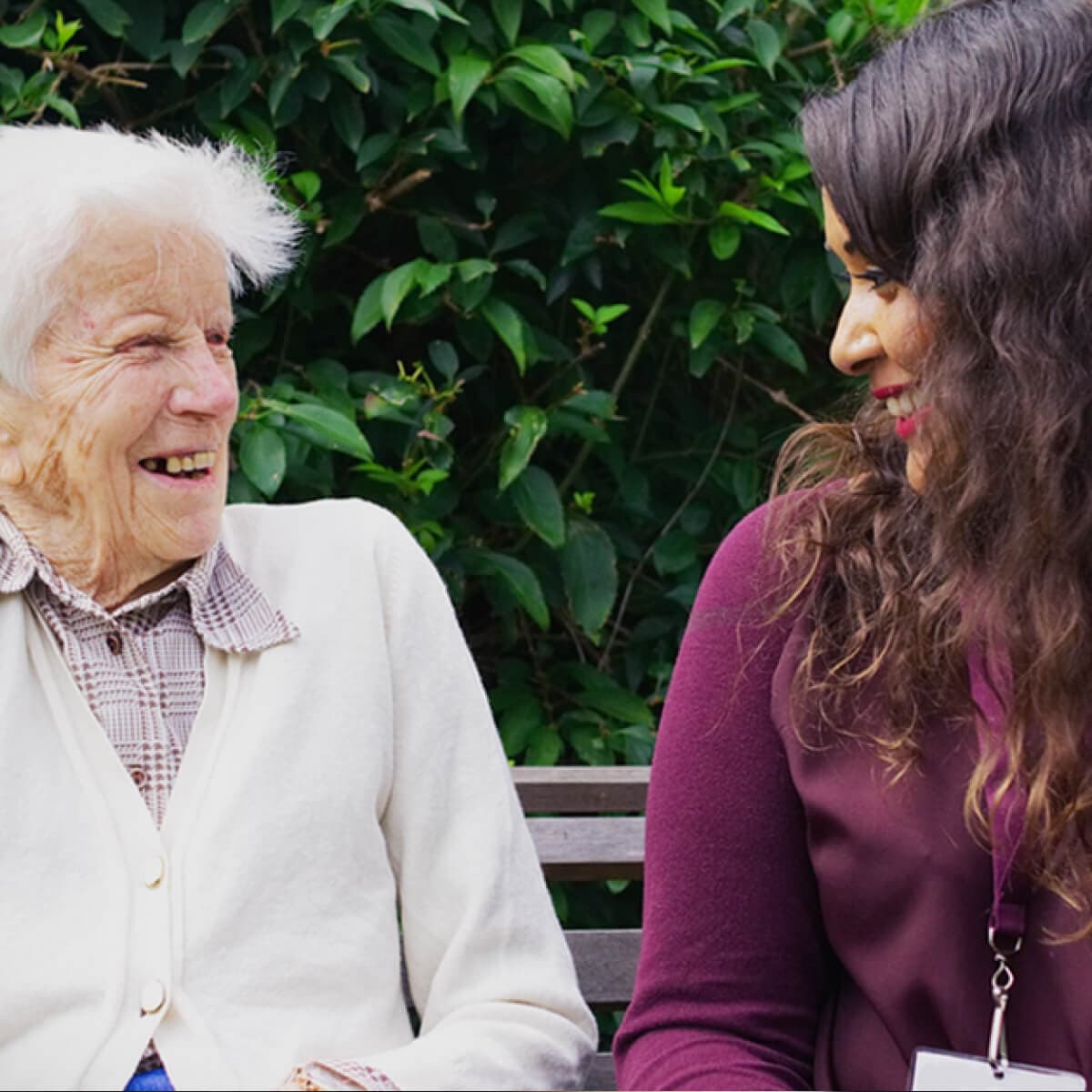 An elderly person and a younger person smile at each other while sitting on a bench in front of greenery. - Home Instead