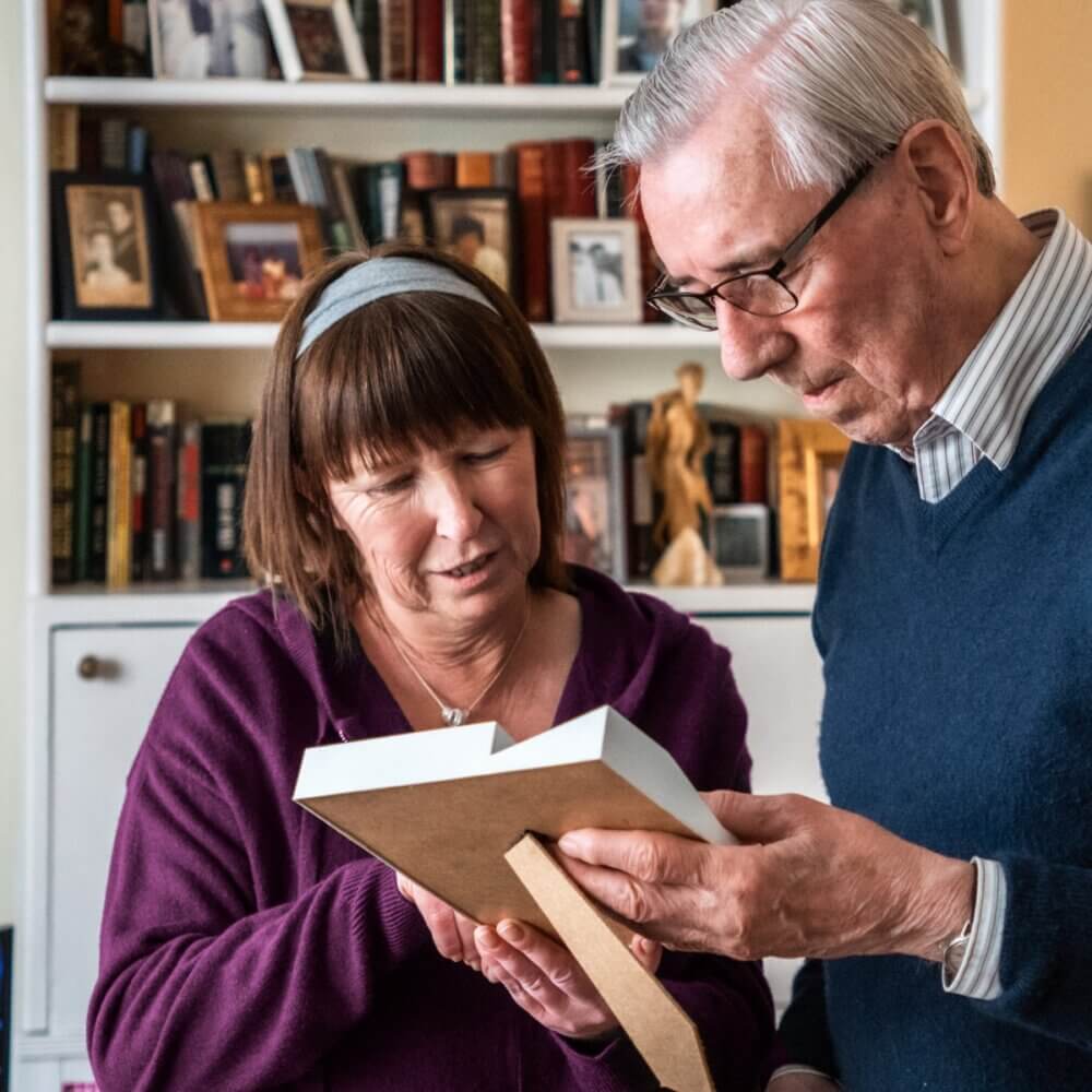 Two elderly individuals looking at a picture frame together in a cozy room with a bookshelf in the background. - Home Instead