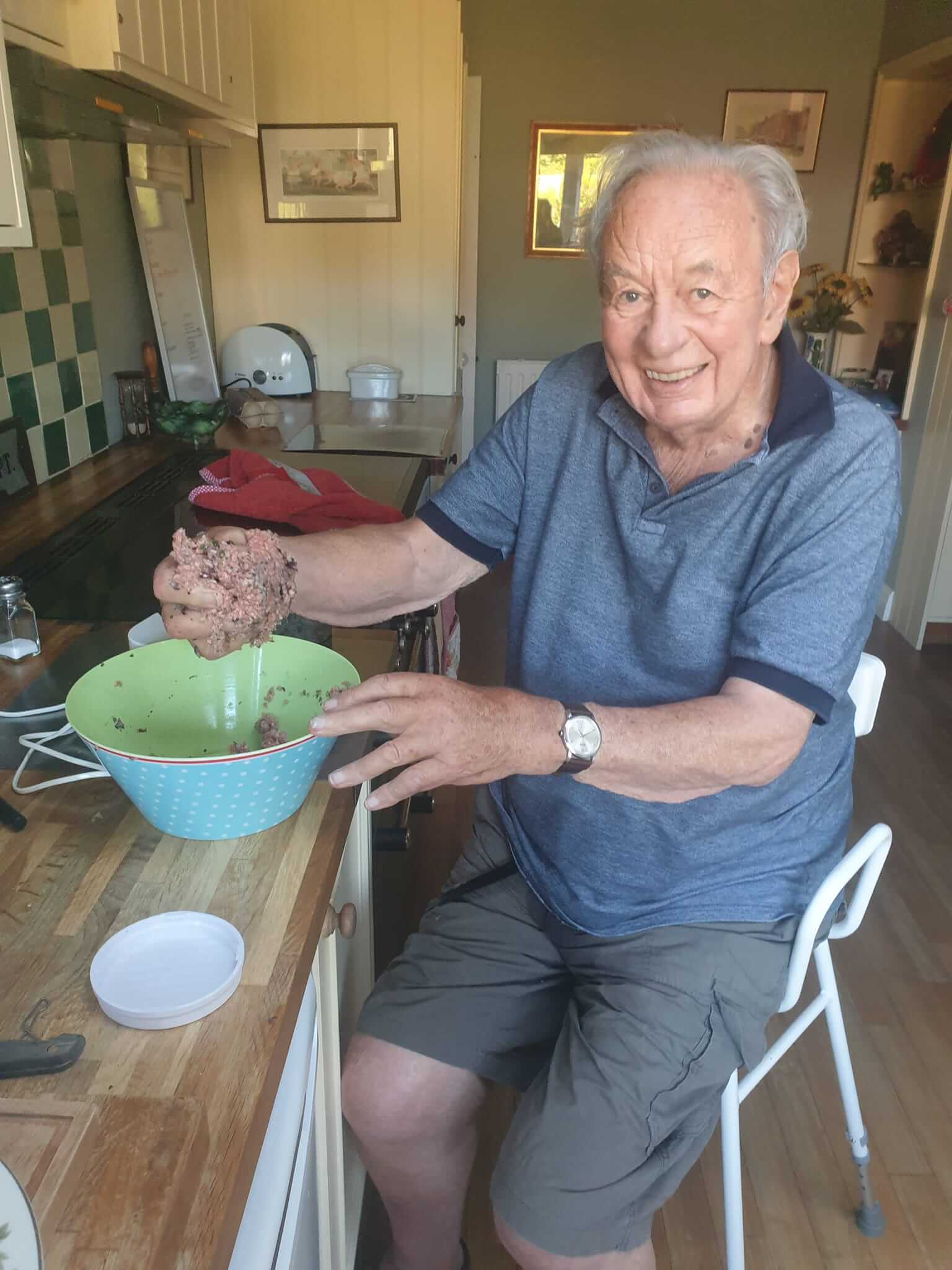 An elderly man, sitting in a kitchen, smiling while mixing ingredients in a bowl. - Home Instead