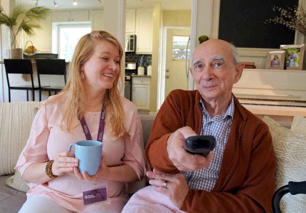 A woman and an elderly man sit on a couch. The man holds a TV remote, and the woman smiles while holding a blue mug. - Home Instead