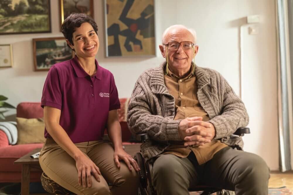 A smiling caregiver in a burgundy shirt sits next to a seated elderly man in a cozy room with artwork on the walls. - Home Instead