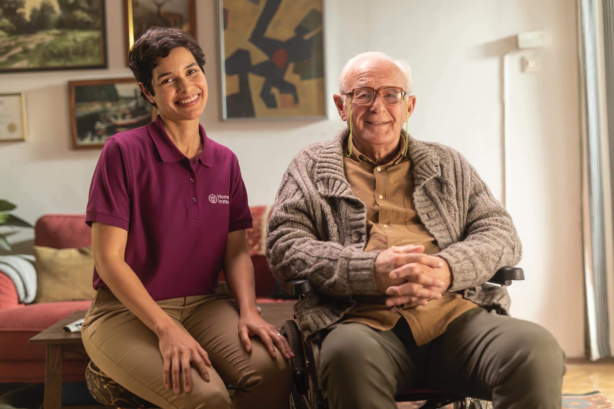 A smiling caregiver in a burgundy shirt sits next to a seated elderly man in a cozy room with artwork on the walls. - Home Instead