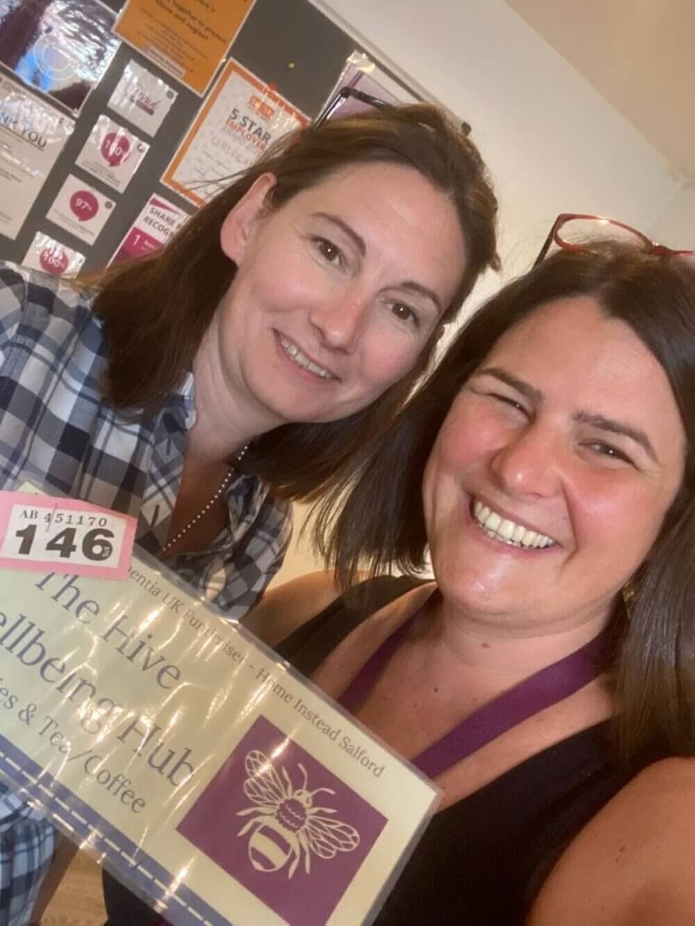 Two women smiling and holding a sign that reads "The Hive Wellbeing Hub - Easters & Tay Coffe." Bulletin board in background. - Home Instead