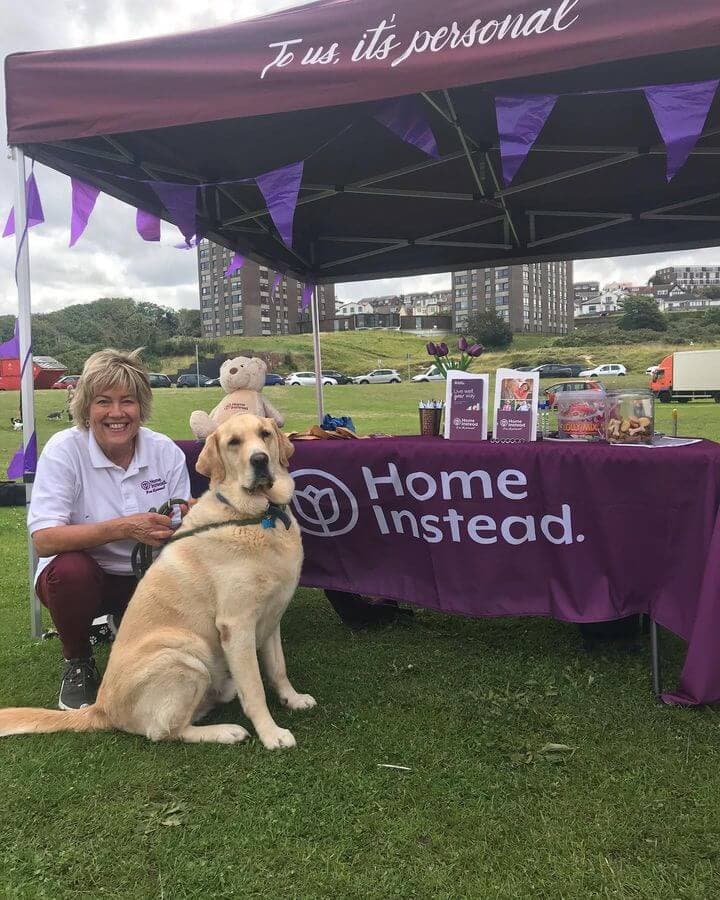A woman and a large dog pose in front of a purple Home Instead table under a tent at an outdoor event. - Home Instead