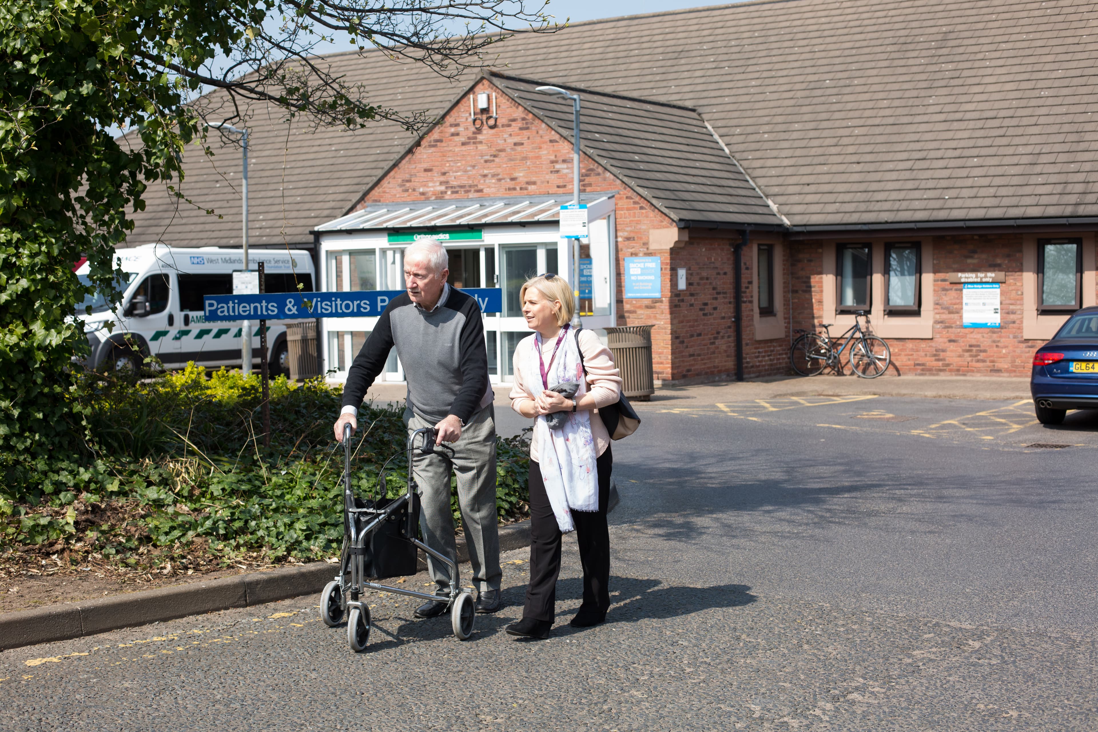 An elderly man using a walker and a woman walk together outside a hospital building with signs for patients and visitors. - Home Instead