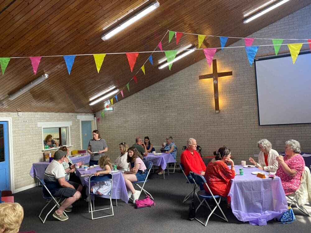 People sitting at tables in a room with colorful bunting, eating and talking, with a cross hanging on the wall. - Home Instead