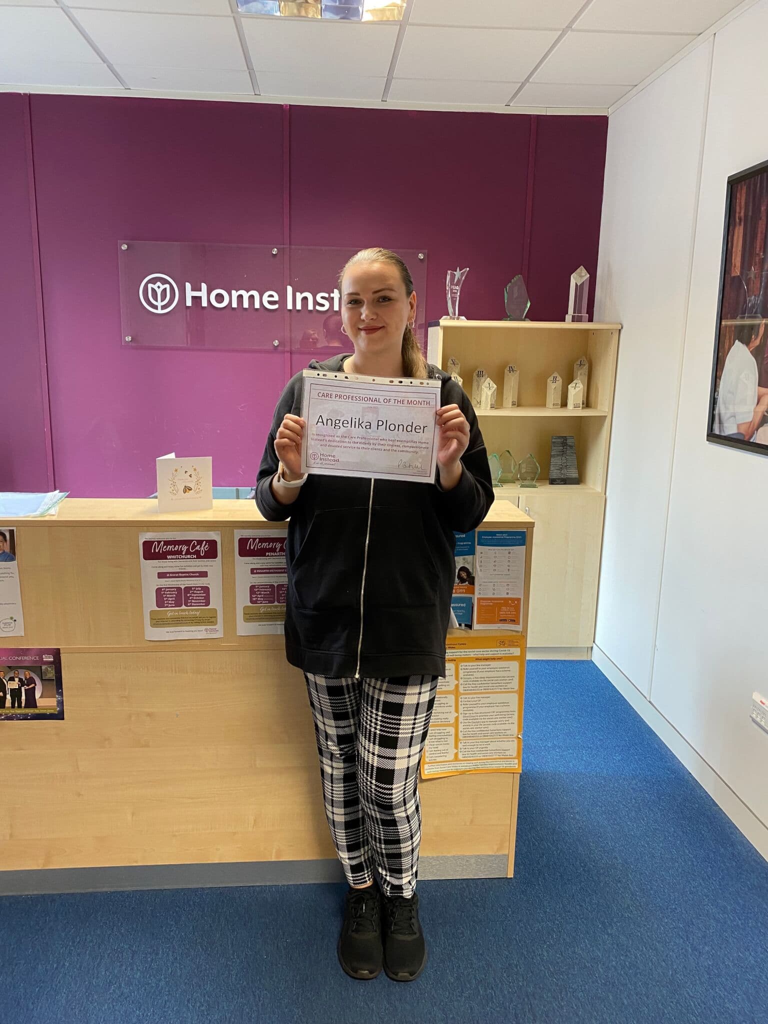 A person holding a certificate and standing in front of a reception desk at "Home Instead" office. - Home Instead