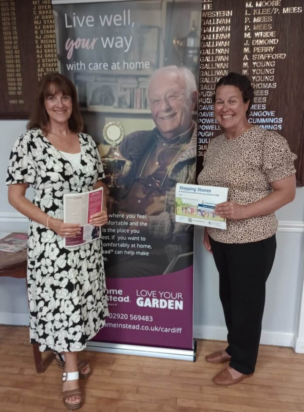 Two women holding brochures, standing in front of a banner with text about home care services. A plaque with names is behind them. - Home Instead
