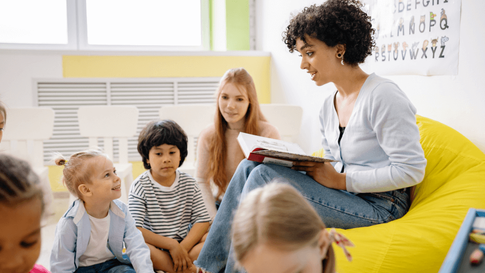 Woman reading a book to a group of children sitting on the floor in a bright, colorful classroom. - Home Instead