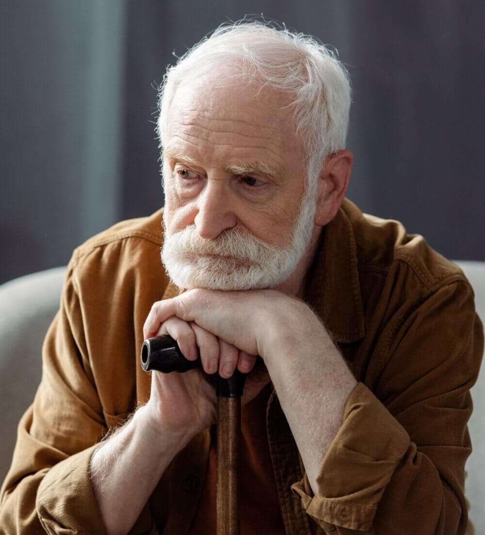 Elderly man with white hair and beard, wearing a brown shirt, resting his chin on a cane, deep in thought. - Home Instead