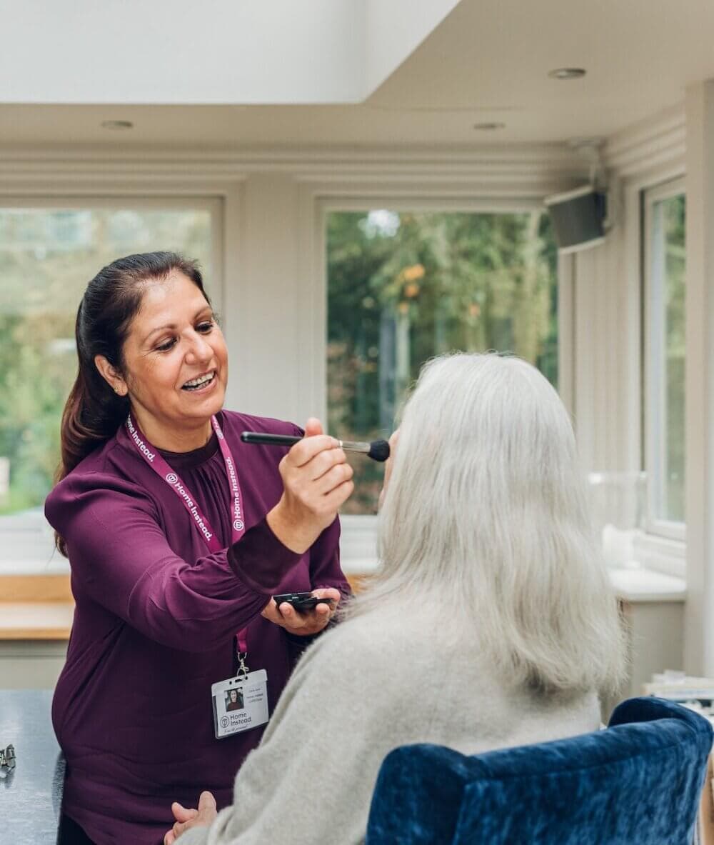 A caregiver in a maroon top applies makeup to an elderly woman with white hair sitting in a chair. - Home Instead