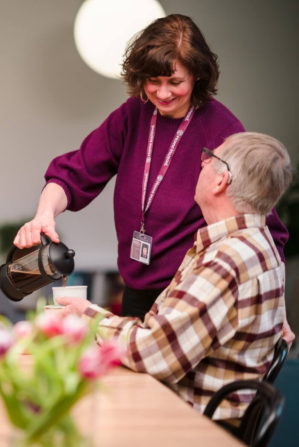 A woman in a purple sweater pours coffee for an older man wearing a plaid shirt at a table with flowers. - Home Instead
