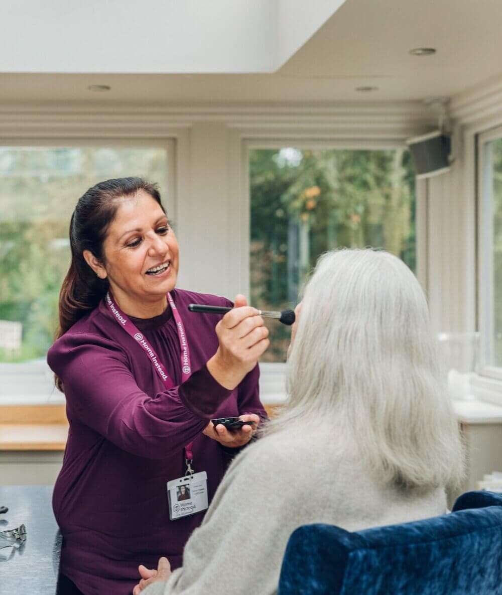 Elderly woman playing a board game at a table with a younger woman wearing a name badge, in a room with framed pictures. - Home Instead