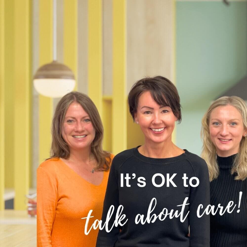 Three women smiling in a brightly lit room with yellow accents. Text reads "It's OK to talk about care!. - Home Instead