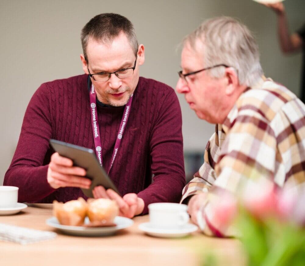 Two men, one younger and one older, sitting at a table with baked goods and cups, looking at a tablet together. - Home Instead