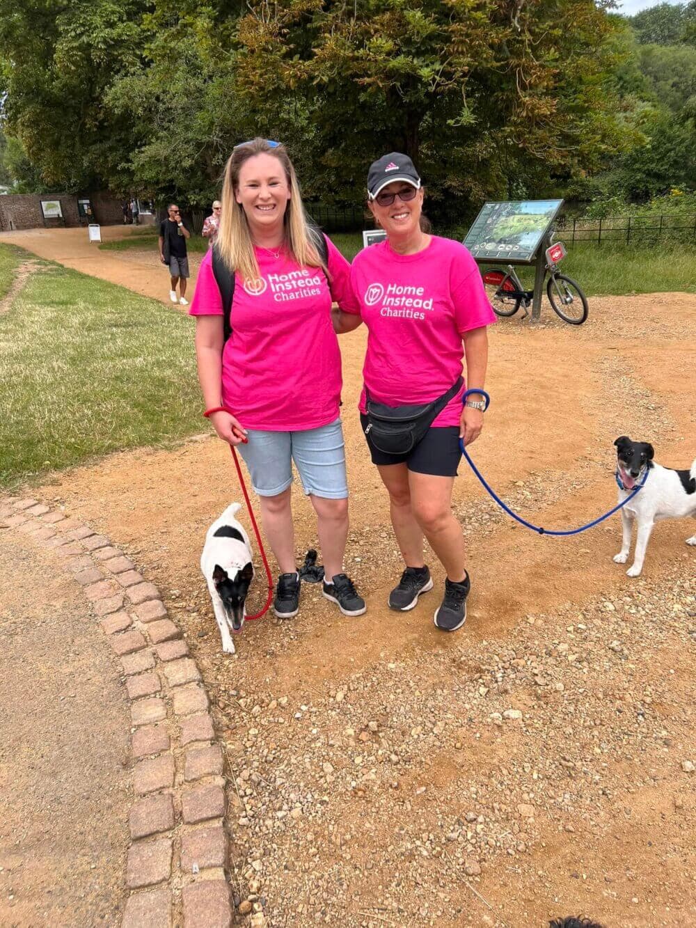 Two women in pink shirts stand on a path with two dogs on leashes, surrounded by greenery and a small crowd. - Home Instead